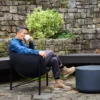 Frame_1_1_1440x Man in a denim shirt sits in a black outdoor chair, sipping from a white mug in a stone-walled garden.