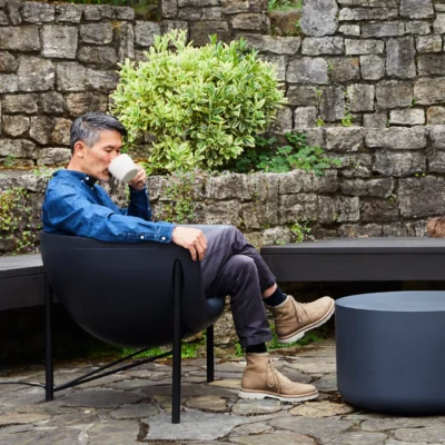 Man in a denim shirt sits in a black outdoor chair, sipping from a white mug in a stone-walled garden.